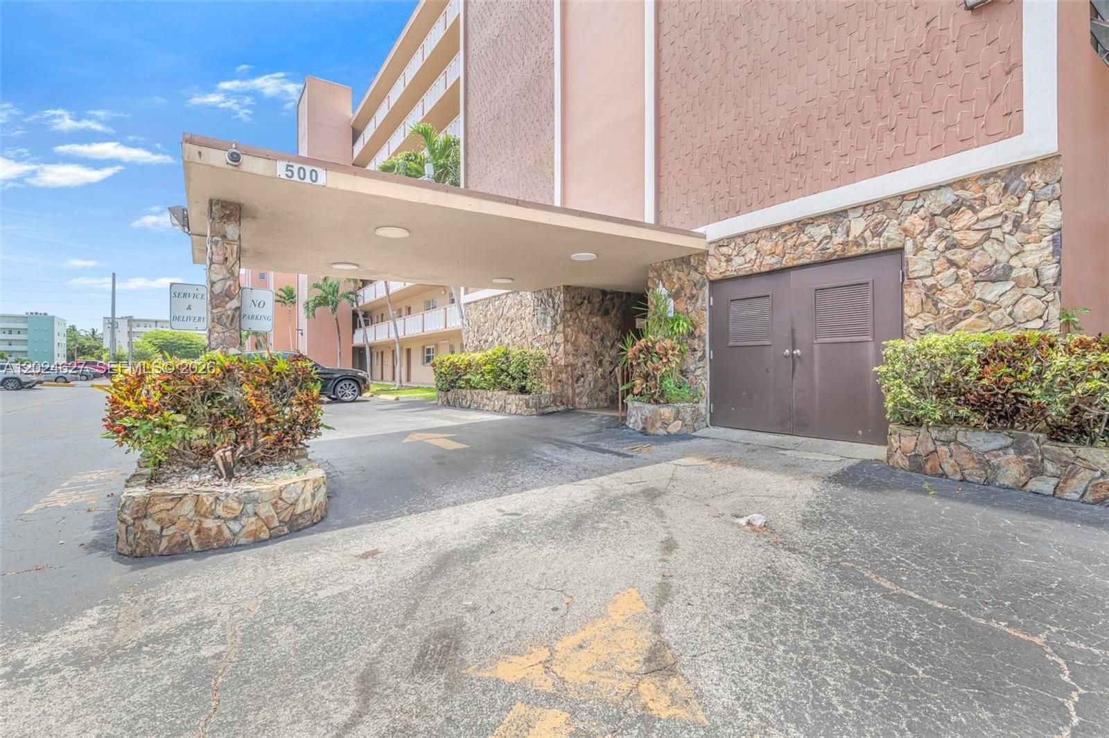 The exterior of a multi-story building features a covered entrance with stone accents and landscaping — 500 NE 12th Ave # 407, Hallandale Beach FL 33009, HALLANDALE BEACH, FL