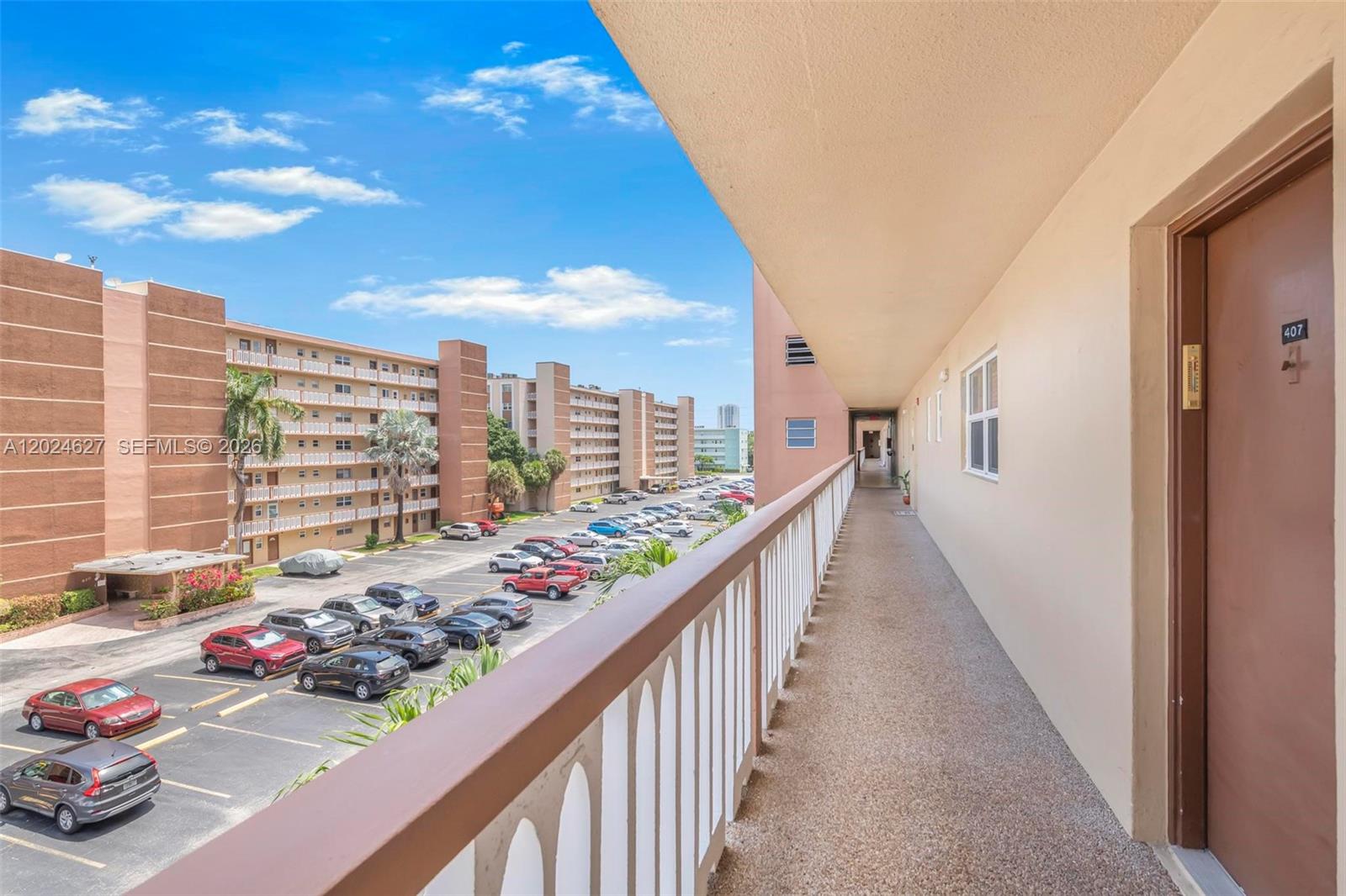An outdoor apartment walkway overlooks a parking lot with multiple cars under a blue sky — 500 NE 12th Ave # 407, Hallandale Beach FL 33009, HALLANDALE BEACH, FL