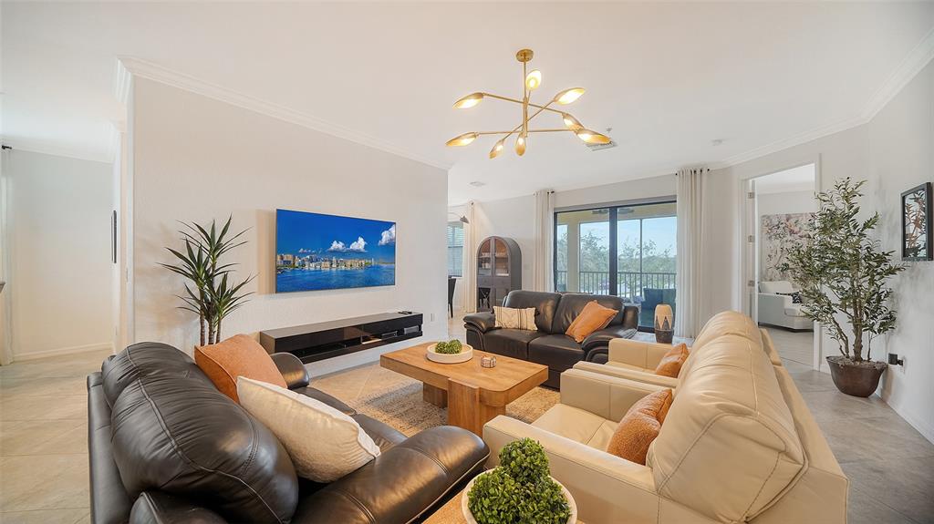 A living room features a wall-mounted television, leather sofas, a wooden coffee table, and chandelier — 6512 GRAND ESTUARY TRAIL UNIT 102, BRADENTON, FL