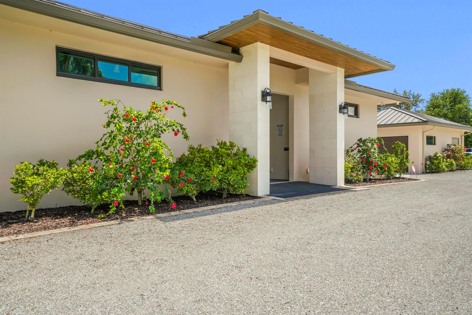 A modern house exterior features a gravel driveway, stone pillars, and hibiscus bushes — 16219 Hollow Tree Lane, Wellington FL 33470, WELLINGTON, FL