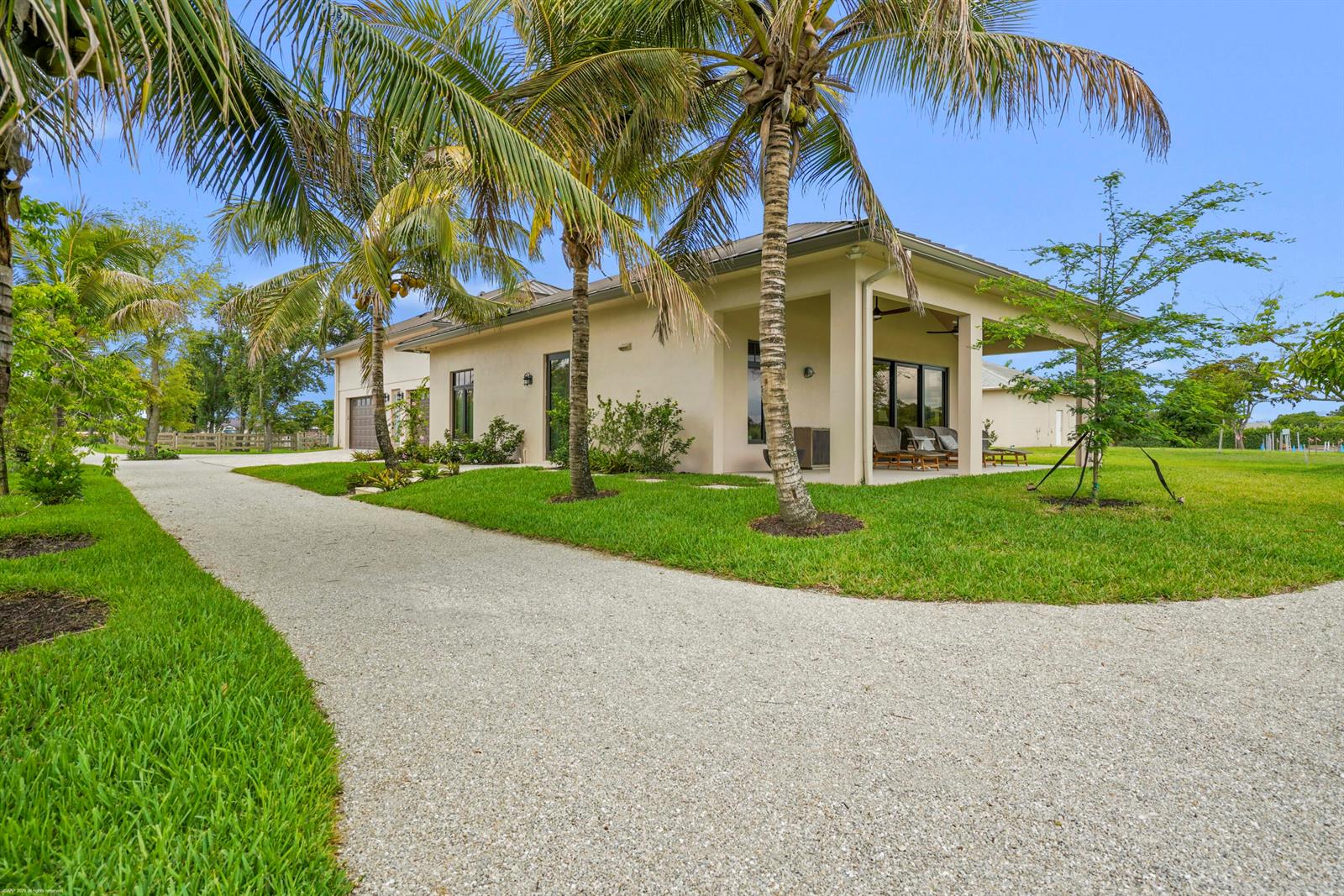 A beige house with a gravel driveway and palm trees under a clear blue sky — 16219 Hollow Tree Lane, Wellington FL 33470, WELLINGTON, FL