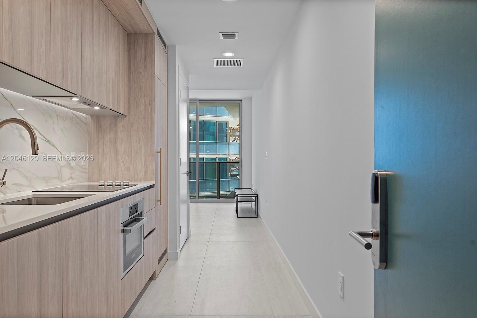 A modern kitchen with light wood cabinetry and marble backsplash leads to a glass balcony door — 77 SE 5th St. # 1905, Miami FL 33131, MIAMI, FL
