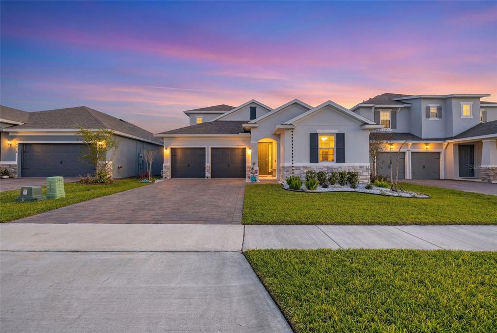 A modern suburban house with a paved driveway, two-car garage, and stone-accented exterior at sunset — 4542 LIONS GATE AVENUE, CLERMONT, FL