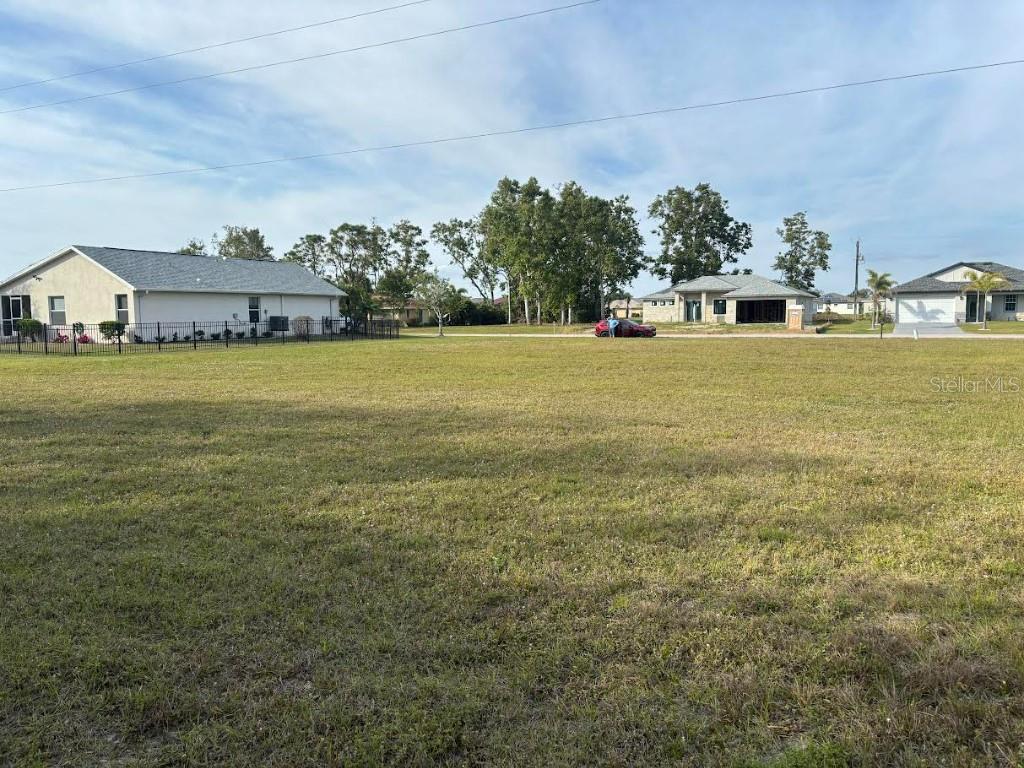 A large, grassy vacant lot is surrounded by residential houses and trees under a blue sky — 25251 DOREDO DRIVE, PUNTA GORDA, FL