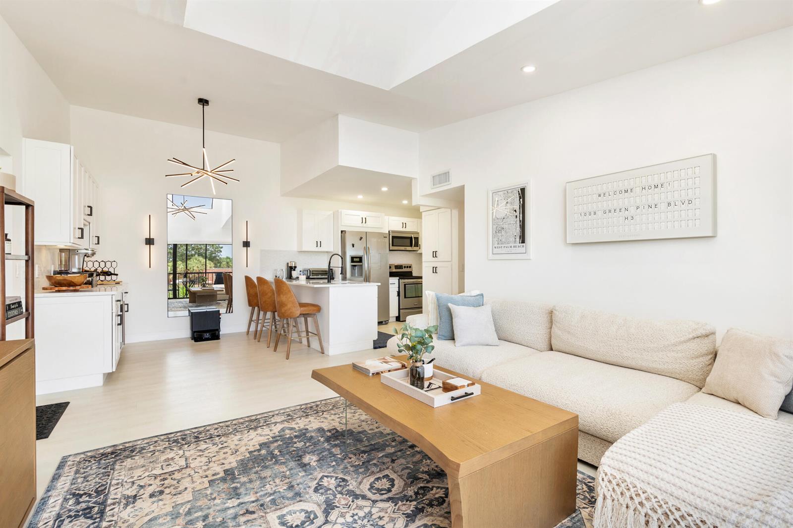 A bright, open-concept living room and kitchen featuring white cabinetry, a sectional sofa, and rugs — 1008 Green Pine Boulevard # H3, West Palm Beach FL 33409, WEST PALM BEACH, FL