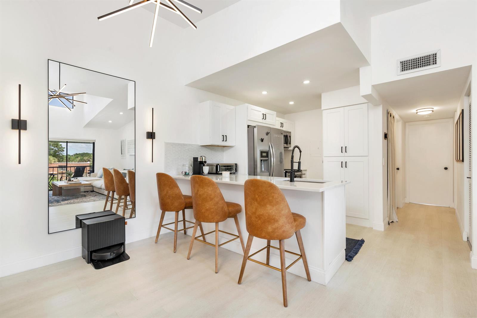 A modern kitchen features white cabinetry, a breakfast bar with leather stools, and a large mirror — 1008 Green Pine Boulevard # H3, West Palm Beach FL 33409, WEST PALM BEACH, FL