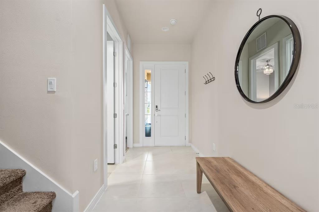 A bright entryway features light tile flooring, a wooden bench, a round mirror, and stairs — 7568 SUNFLOWER CIRCLE, KISSIMMEE, FL