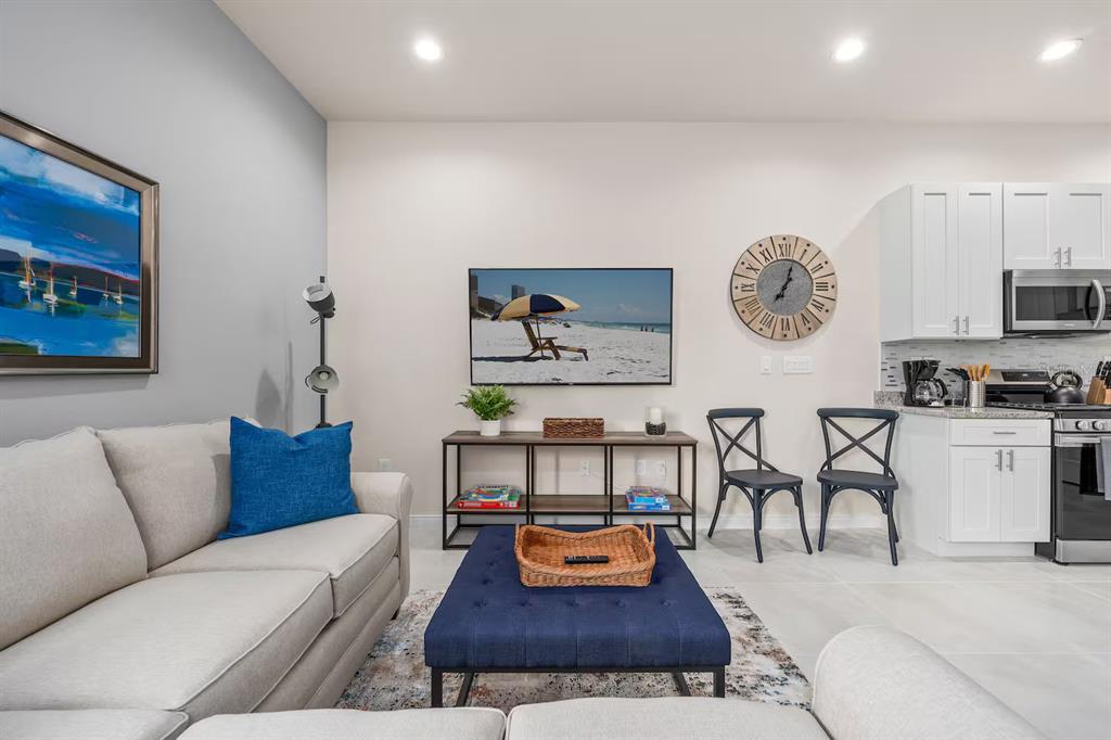 A living room with a beige sofa, blue ottoman, television, and an adjacent kitchen area — 7568 SUNFLOWER CIRCLE, KISSIMMEE, FL