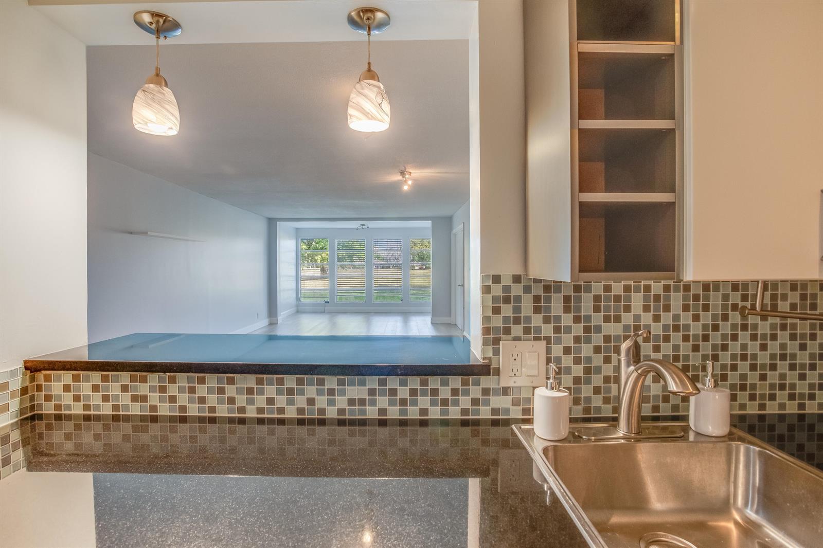 A kitchen sink with dark countertops and a tiled backsplash overlooks an open living area — 2651 S Course Drive # 109, Pompano Beach FL 33069, POMPANO BEACH, FL