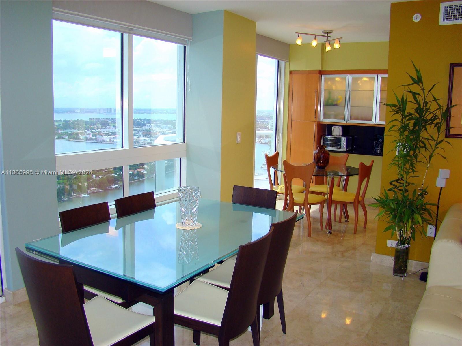 A dining area with two glass-topped tables, modern chairs, and floor-to-ceiling windows overlooking water — 6365 COLLINS AV # 3407, Miami Beach FL 33141, MIAMI BEACH, FL