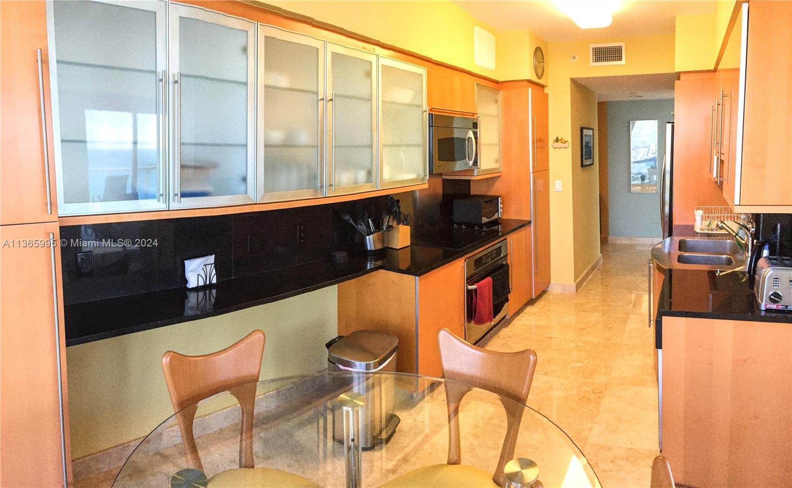 A kitchen features light wood cabinets, black countertops, stainless steel appliances, and a glass table — 6365 COLLINS AV # 3407, Miami Beach FL 33141, MIAMI BEACH, FL