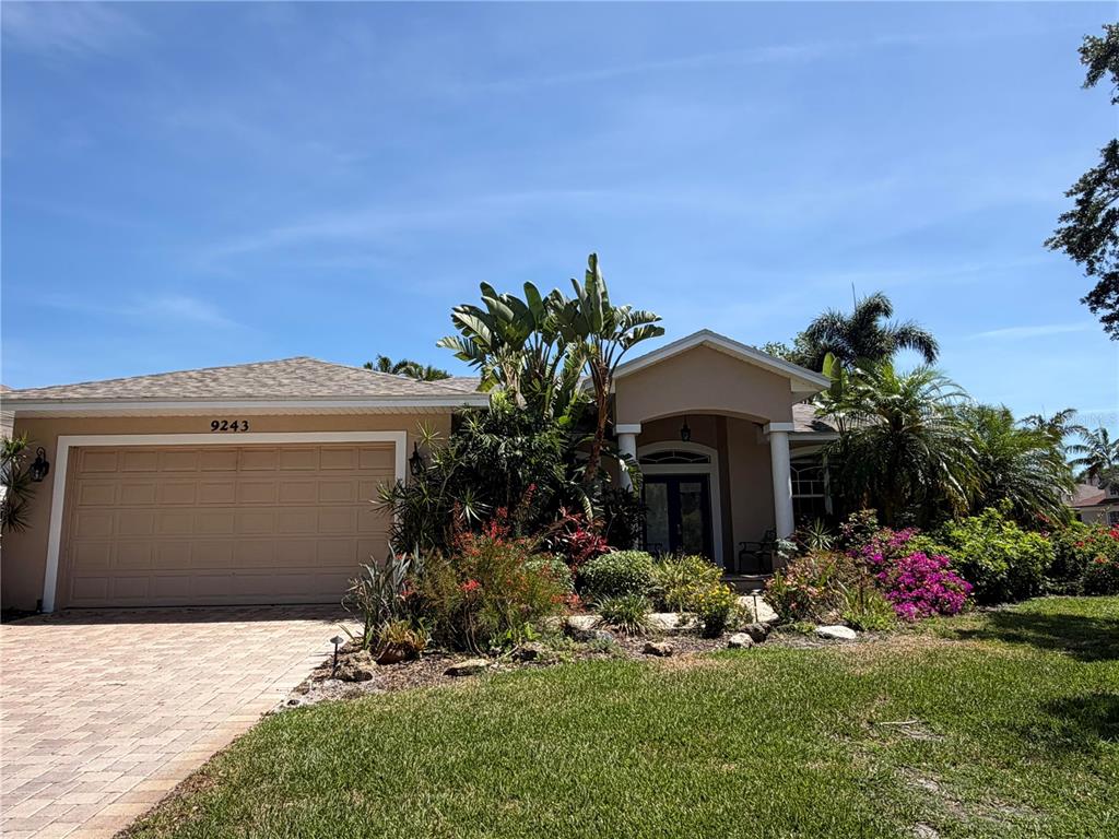 A single-story house with a tan exterior, a two-car garage, and lush tropical landscaping — 9243 13TH AVENUE CIRCLE NW, BRADENTON, FL