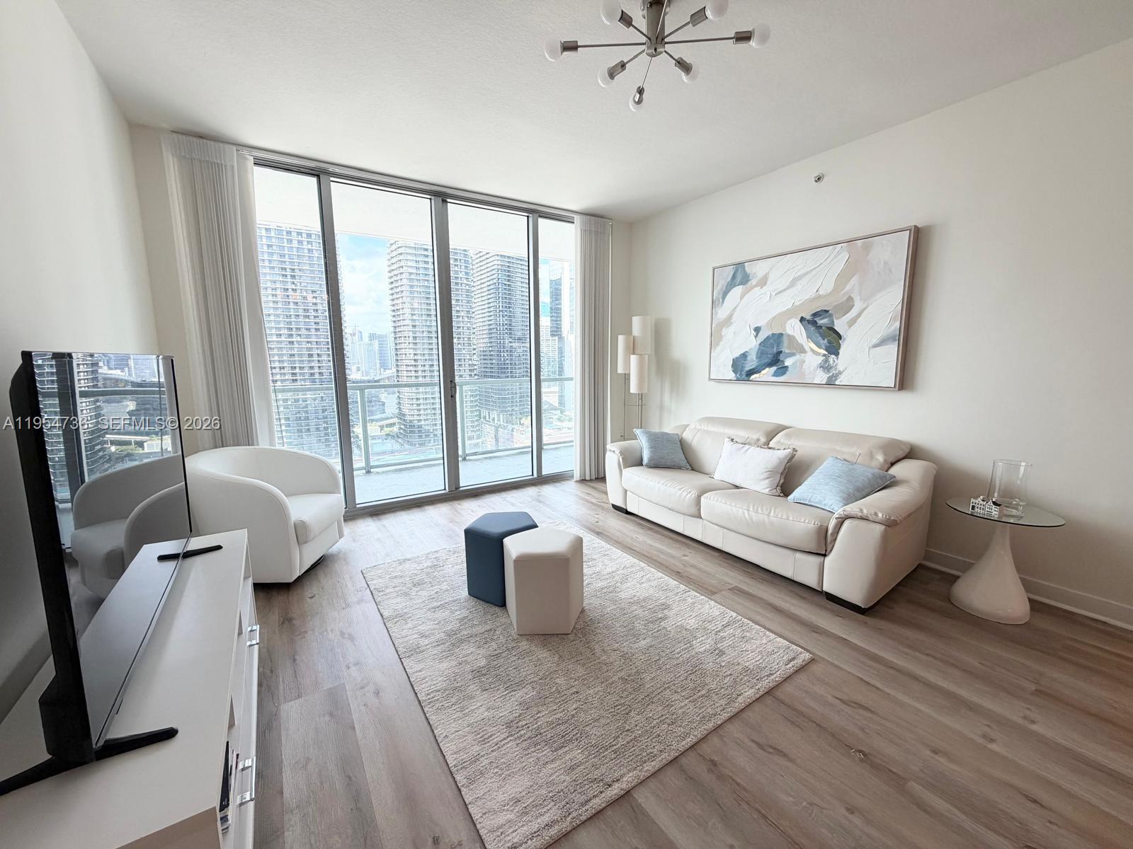 A modern living room features a white sofa, a television, and floor-to-ceiling windows overlooking skyscrapers — 1111 SW 1 AV, Miami FL 33130, MIAMI, FL