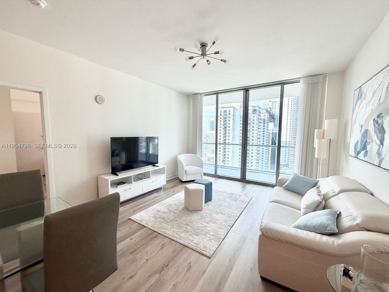 A modern living room features a white sofa, television console, and floor-to-ceiling windows overlooking city buildings — 1111 SW 1 AV, Miami FL 33130, MIAMI, FL