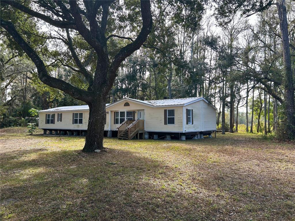 A light-colored manufactured home sits on a pier foundation among trees in a grassy yard — 9867 JEROME DRIVE, NEW PORT RICHEY, FL