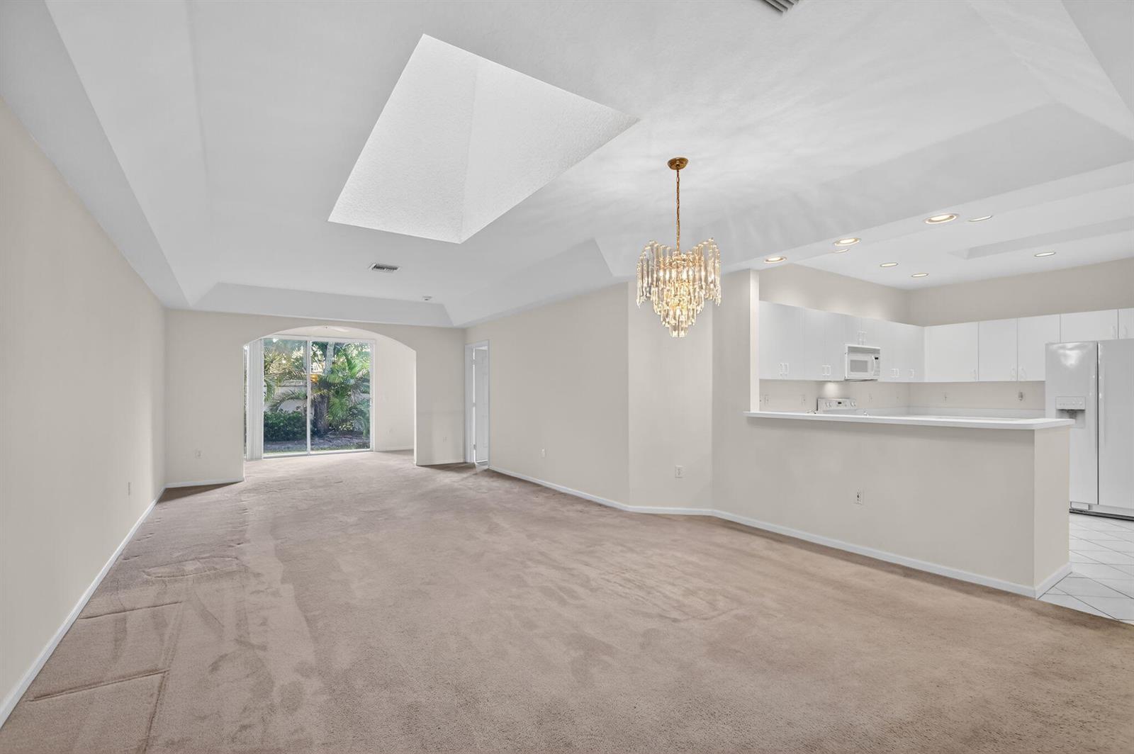 An open-concept living area with beige carpeting, a crystal chandelier, and an adjacent white kitchen — 12574 Crystal Pointe Drive # C, Boynton Beach FL 33437, BOYNTON BEACH, FL