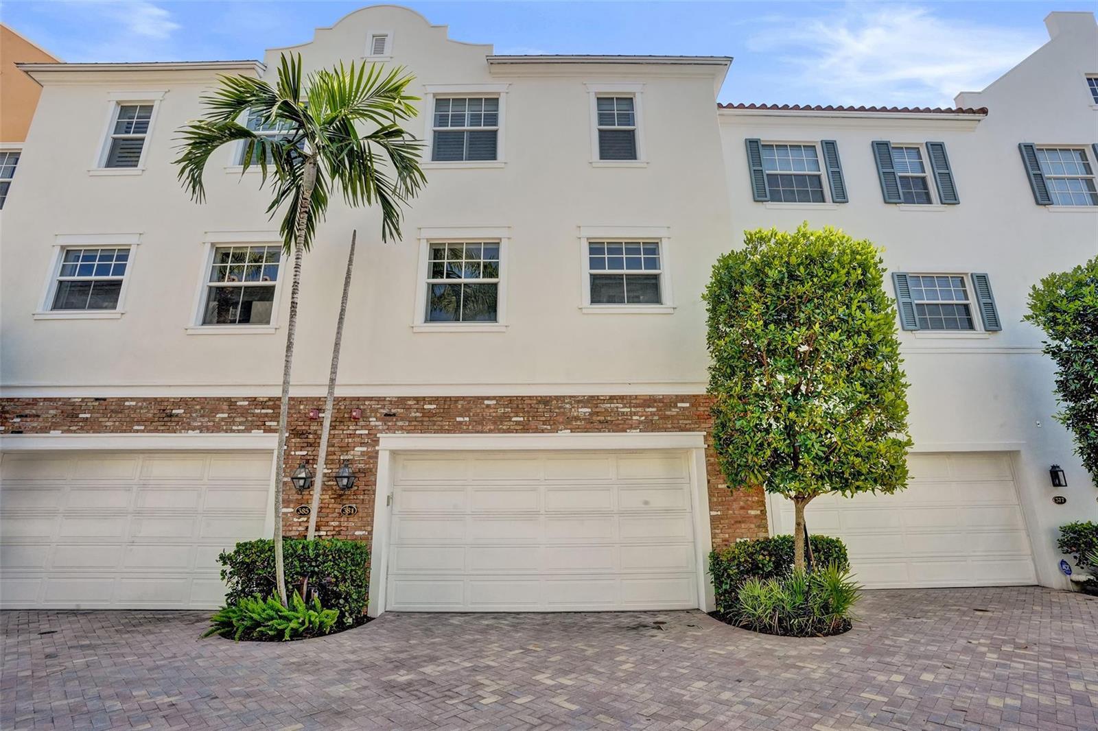 A three-story townhouse exterior features white stucco walls, brick accents, and three garage doors — 381 E Cannery Row Circle, Delray Beach FL 33444, DELRAY BEACH, FL