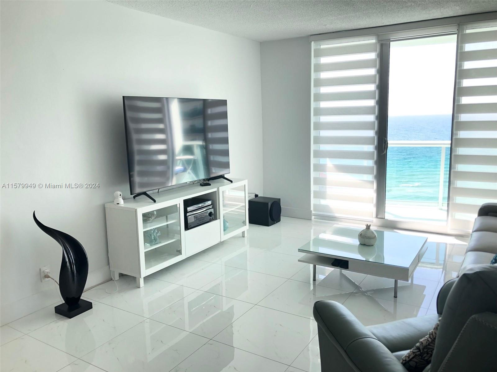 A modern living room features white tile floors, a television console, and ocean views — 3901 S Ocean Dr # 14M, Hollywood FL 33019, HOLLYWOOD, FL