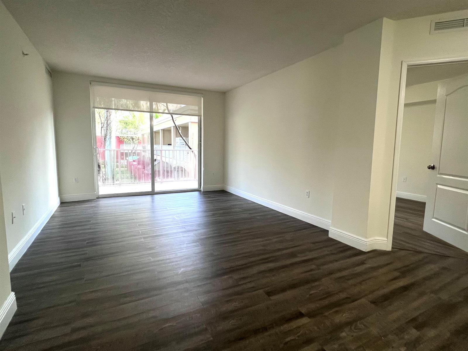 An empty living room features dark wood-look flooring, white walls, and a sliding glass door — 18335 NW 68 Avenue # 406, Hialeah FL 33015, HIALEAH, FL