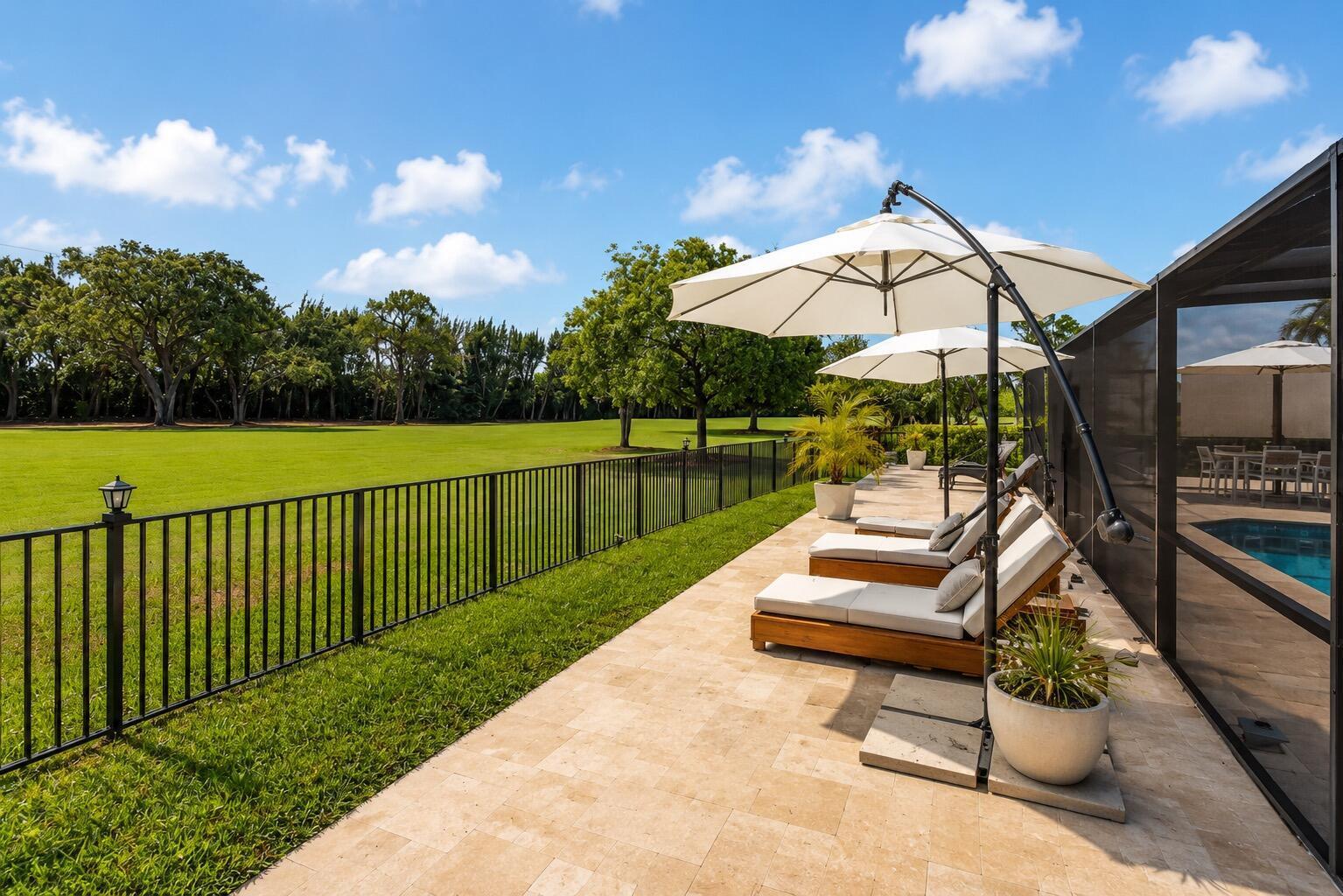 A patio with lounge chairs and umbrellas overlooks a large grassy lawn and black fence — 10710 Boca Woods Lane, Boca Raton FL 33428, BOCA RATON, FL
