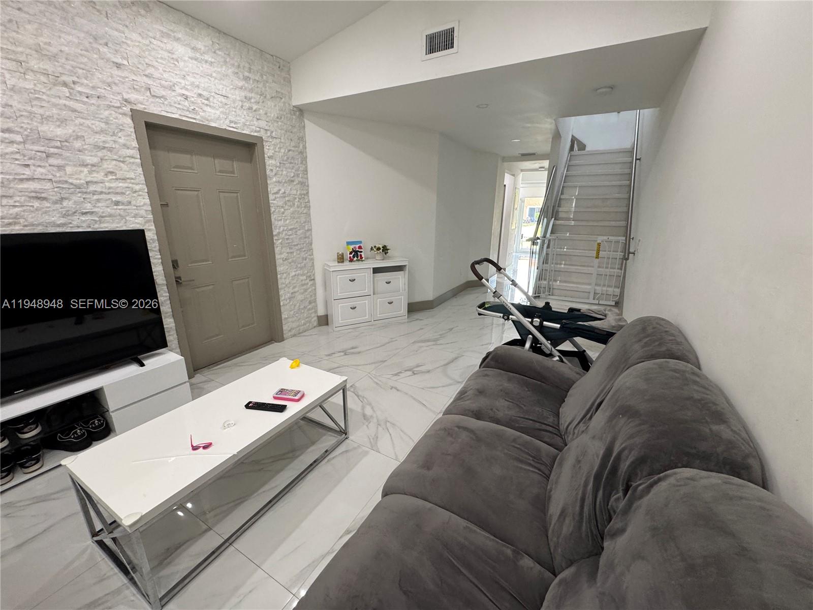 A living room features a stone accent wall, white tile flooring, and a gray sofa — 4957 Tradewinds Ter # 704, Dania Beach FL 33312, DANIA BEACH, FL