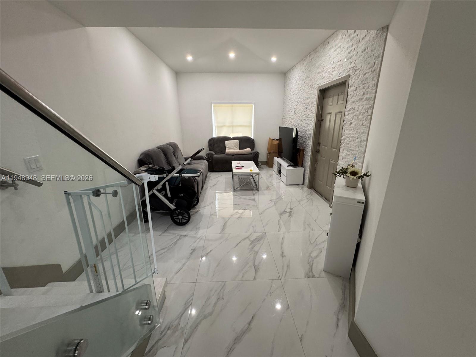 A living room features white marble-look tile flooring, a textured stone wall, and a staircase — 4957 Tradewinds Ter # 704, Dania Beach FL 33312, DANIA BEACH, FL