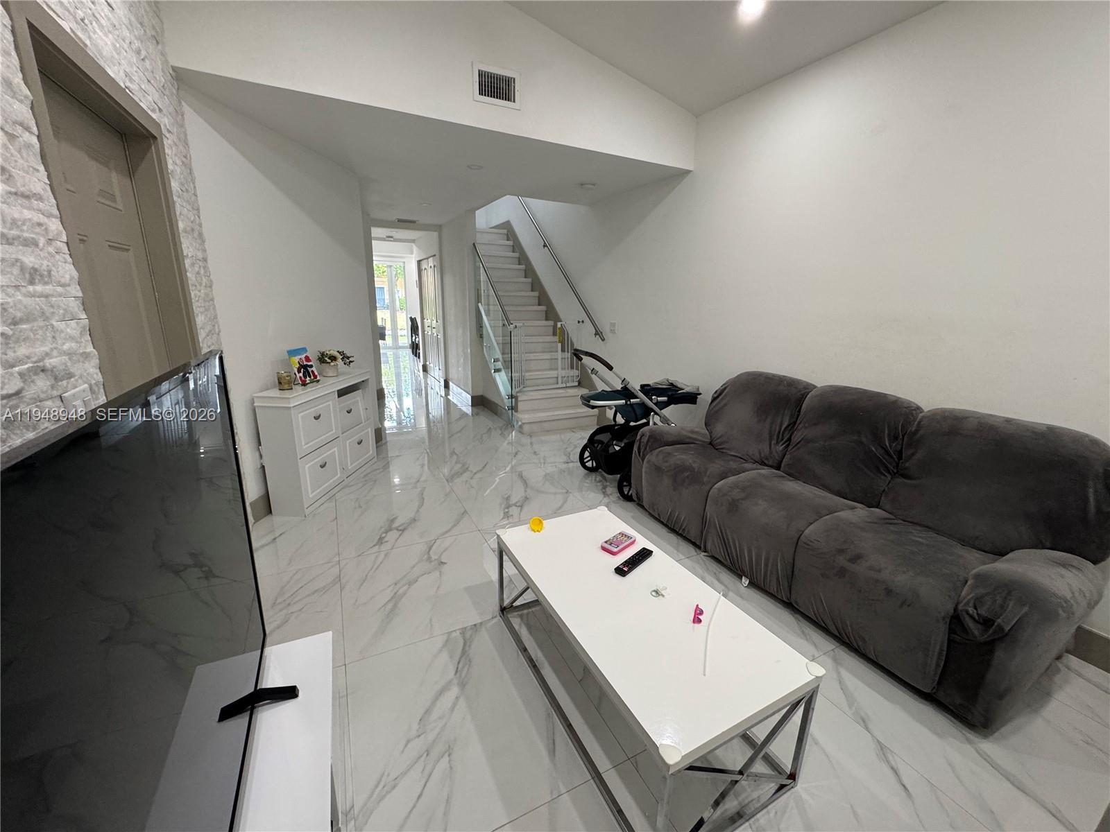 A living room features white marble-style tile flooring, a grey sofa, and a staircase — 4957 Tradewinds Ter # 704, Dania Beach FL 33312, DANIA BEACH, FL