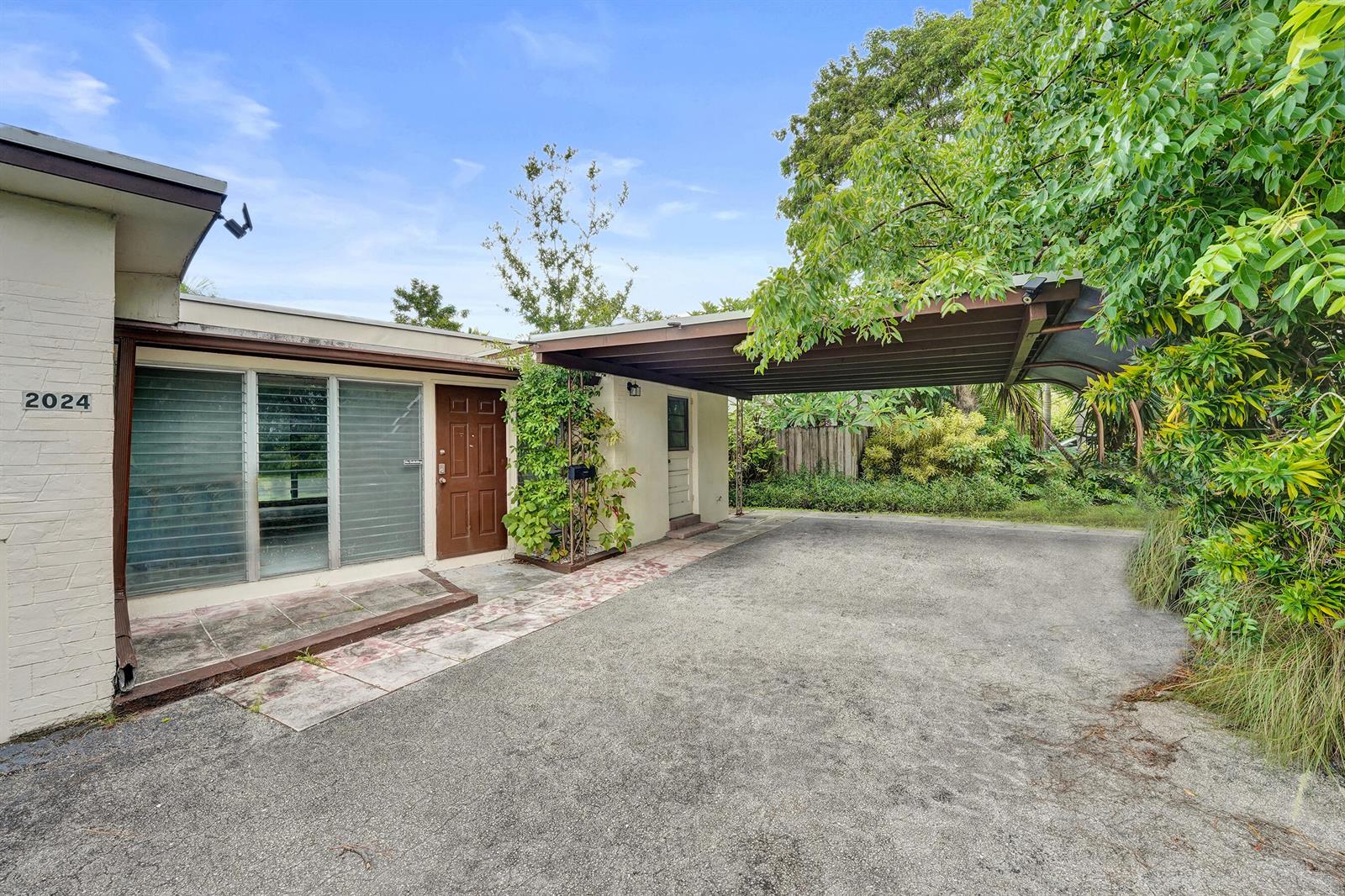 A single-story house exterior features a brown front door, jalousie windows, and a metal carport — 2024 NE 26th Drive, Wilton Manors FL 33306, WILTON MANORS, FL