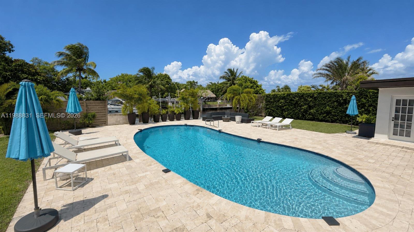 A backyard features a swimming pool, lounge chairs, umbrellas, and tropical landscaping under a blue sky — 1805 Keystone Blvd, North Miami FL 33181, NORTH MIAMI, FL