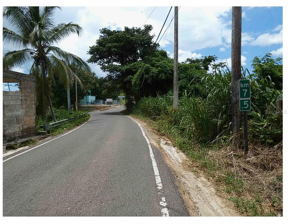 A paved road winds through a rural area with a kilometer marker sign on the right — SR 108 KM 7.4 RIO CAÑAS WD UNIT C-1, ANASCO, PR