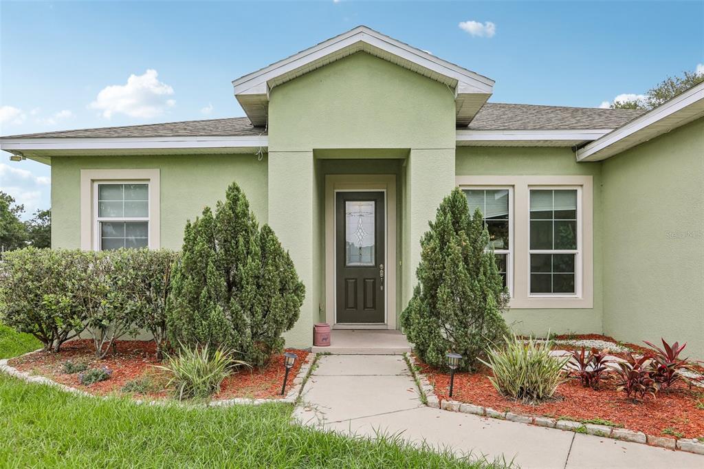 A light green house exterior features a front door, walkway, and landscaped garden beds — 9876 PANGOLA LOOP, LAND O LAKES, FL