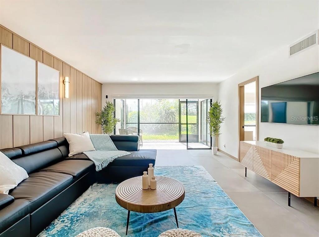 A modern living room features a black sectional sofa, wood-paneled wall, and sliding glass doors — 5532 PUERTA DEL SOL BOULEVARD S UNIT 136, ST PETERSBURG, FL