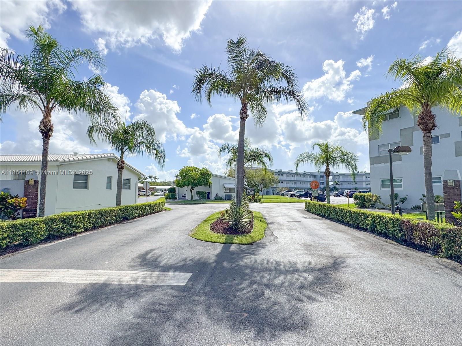 A paved driveway leads between two apartment buildings with palm trees and manicured hedges — 2300 Park Ln # 105, Hollywood FL 33021, HOLLYWOOD, FL