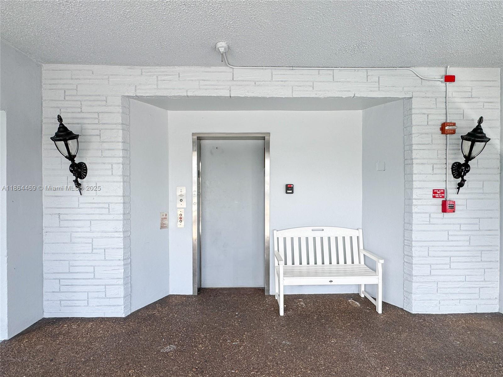 An elevator entrance features a white bench, brick-textured walls, and two black wall-mounted lamps — 2300 Park Ln # 105, Hollywood FL 33021, HOLLYWOOD, FL