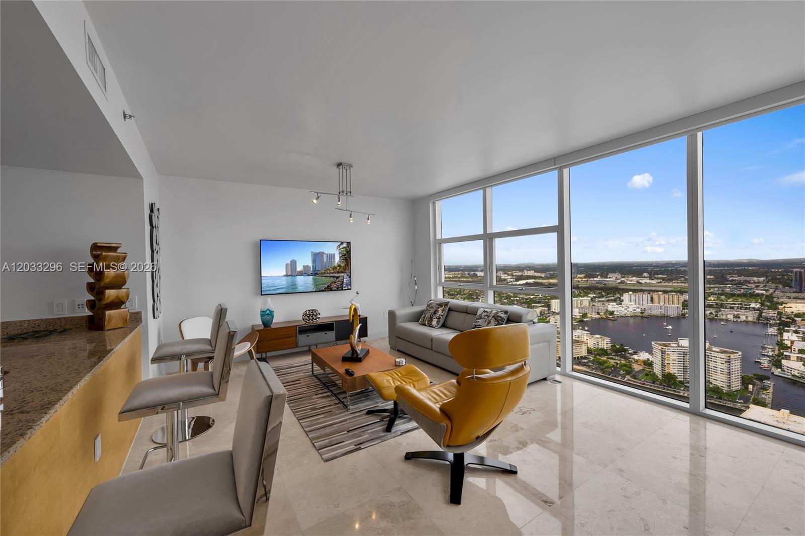 A modern living room features floor-to-ceiling windows overlooking a city skyline and waterfront — 1830 S Ocean Dr # 4807, Hallandale Beach FL 33009, HALLANDALE BEACH, FL