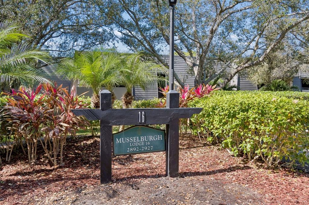 A sign for Musselburgh Lodge 16 stands in a garden bed with shrubs and trees — 36750 US HIGHWAY 19 N UNIT 16210, PALM HARBOR, FL