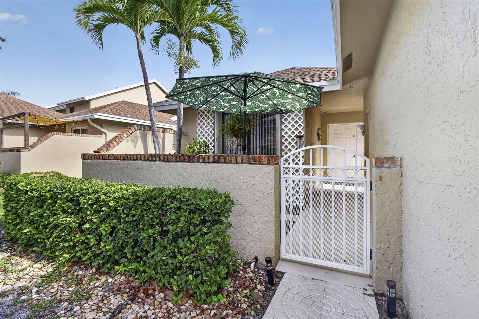 A white gate leads to a patio area with a green umbrella and palm tree — 1101 Maplewood Drive, FL 33415, FL