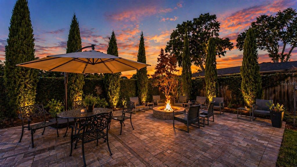 A paved outdoor patio features a fire pit, dining area with umbrella, and tall cypress trees — 906 CRESTVIEW DRIVE, AUBURNDALE, FL