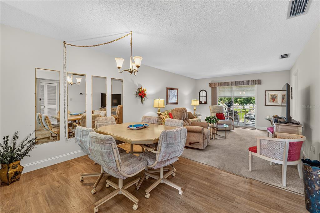 A dining area with a square table and chairs sits adjacent to a carpeted living room — 4801 AIRPORT ROAD UNIT 119, ZEPHYRHILLS, FL