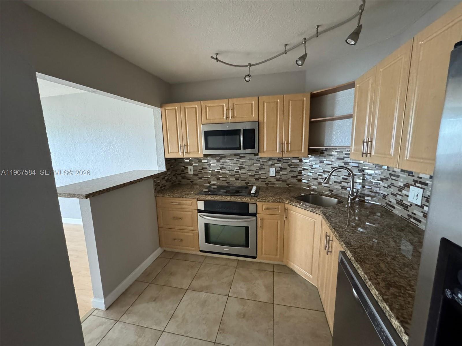 A kitchen features light wood cabinets, granite countertops, a tile backsplash, and stainless steel appliances — 3301 Spanish Moss Ter # 604, Lauderhill FL 33319, LAUDERHILL, FL