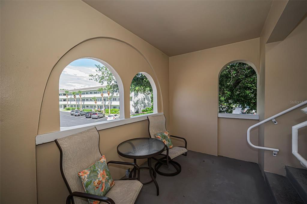 A covered balcony features two chairs, a small table, and arched openings overlooking a parking lot — 2384 TAHITIAN LANE UNIT 22, CLEARWATER, FL