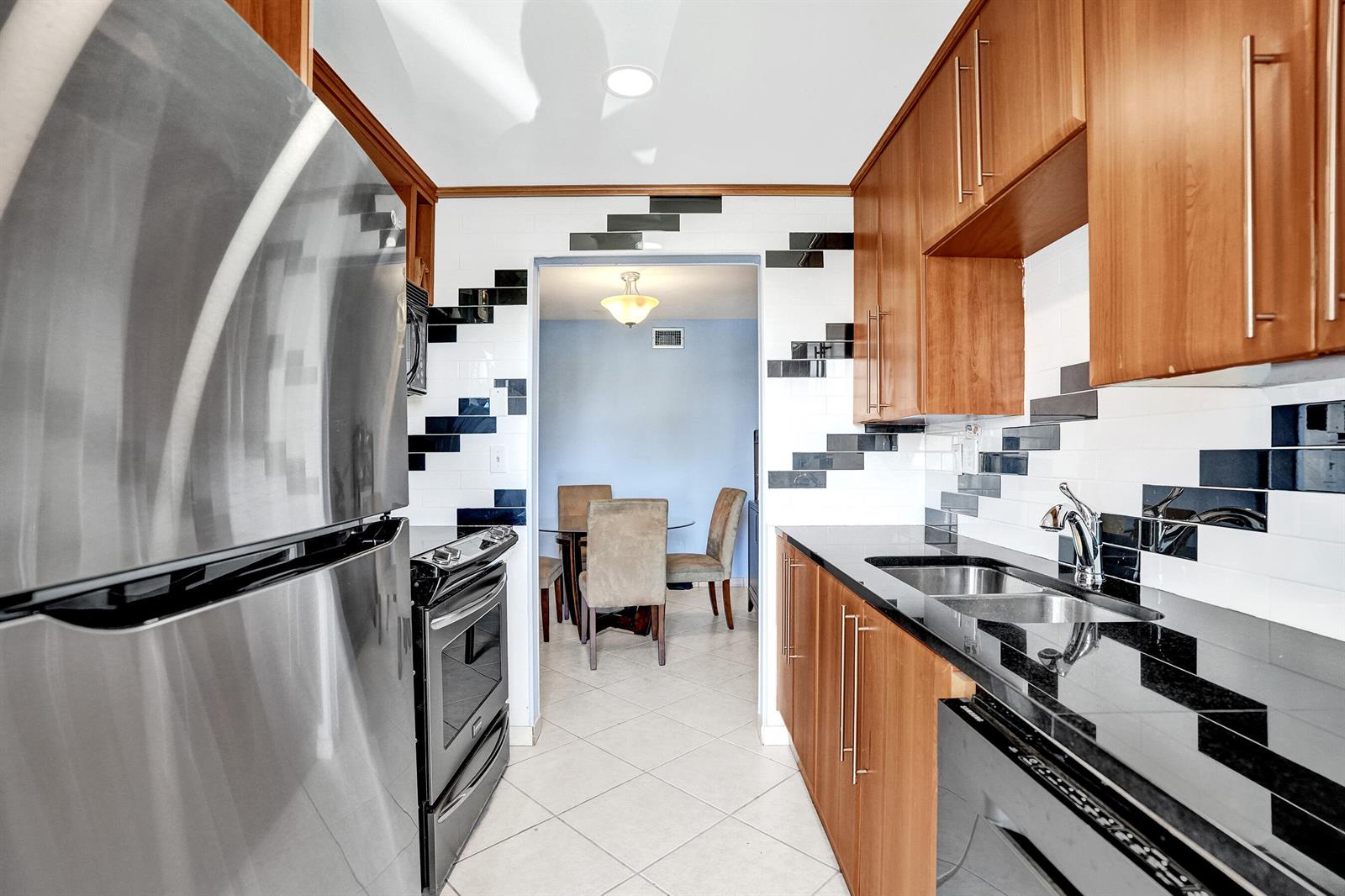 A galley kitchen features wood cabinets, black countertops, and a unique black and white tile backsplash — 930 SE 9th Avenue # 11, Pompano Beach FL 33060, POMPANO BEACH, FL