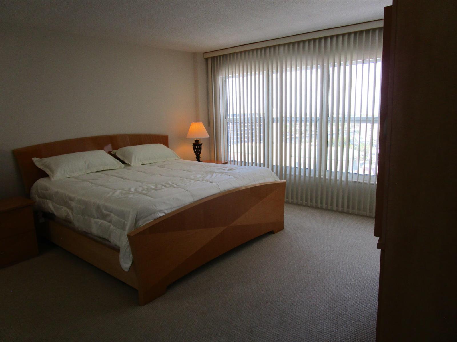 A bedroom with a wooden bed frame, white bedding, beige carpet, and vertical window blinds — 405 N Ocean Boulevard # 1225, Pompano Beach FL 33062, POMPANO BEACH, FL