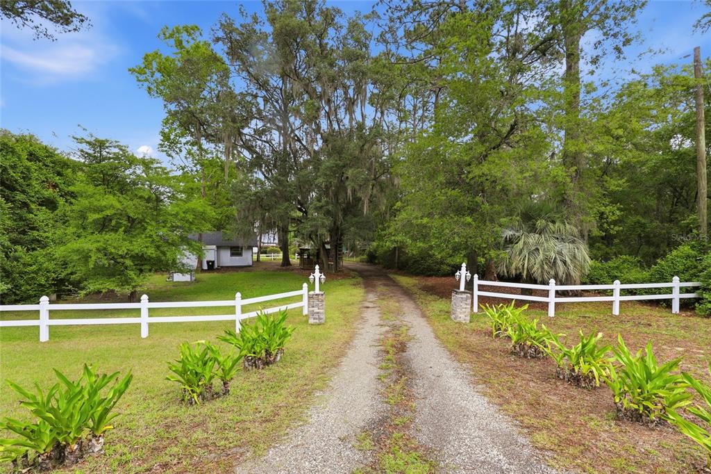 A gravel driveway leads through a white fence toward a house shaded by large trees — 5419 COUNTY ROAD 352, KEYSTONE HEIGHTS, FL