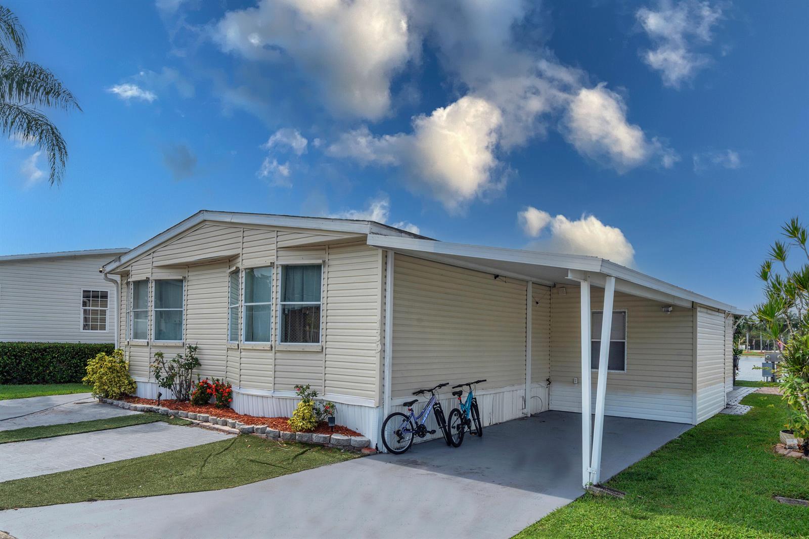 A beige manufactured home features a carport, a paved driveway, and a small landscaped garden — 139 Plantation Boulevard, FL 33467, FL