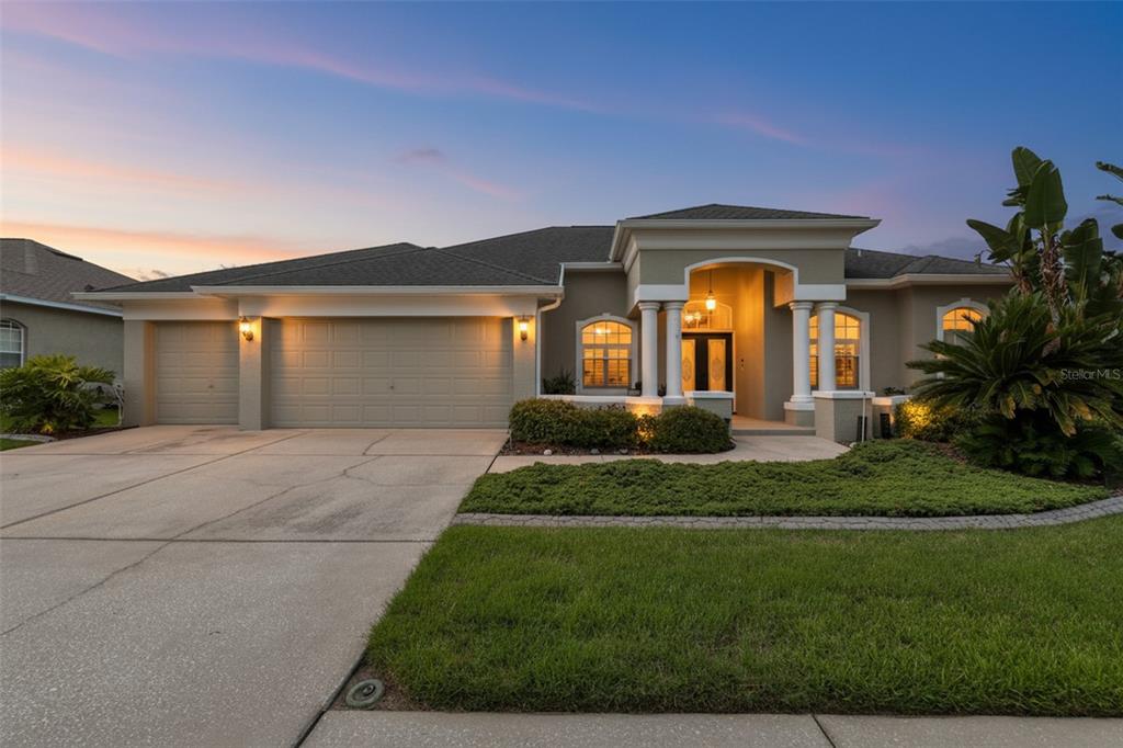 A single-story house with a three-car garage, arched entryway, and landscaped front yard at dusk — 7521 MELOGOLD CIRCLE, LAND O LAKES, FL