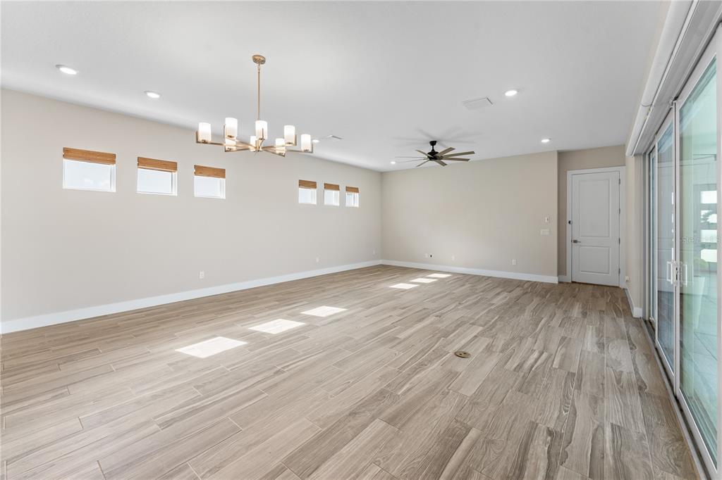 A spacious, empty room features light wood-look tile flooring, a modern chandelier, and sliding glass doors — 8249 IVY STARK BOULEVARD, WESLEY CHAPEL, FL