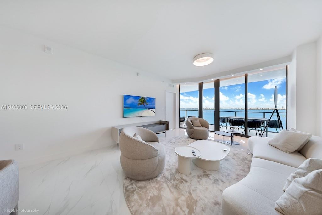 A modern living room features white marble floors, curved furniture, and floor-to-ceiling windows overlooking water — 700 NE 24 St # 1403, Miami FL 33137, MIAMI, FL