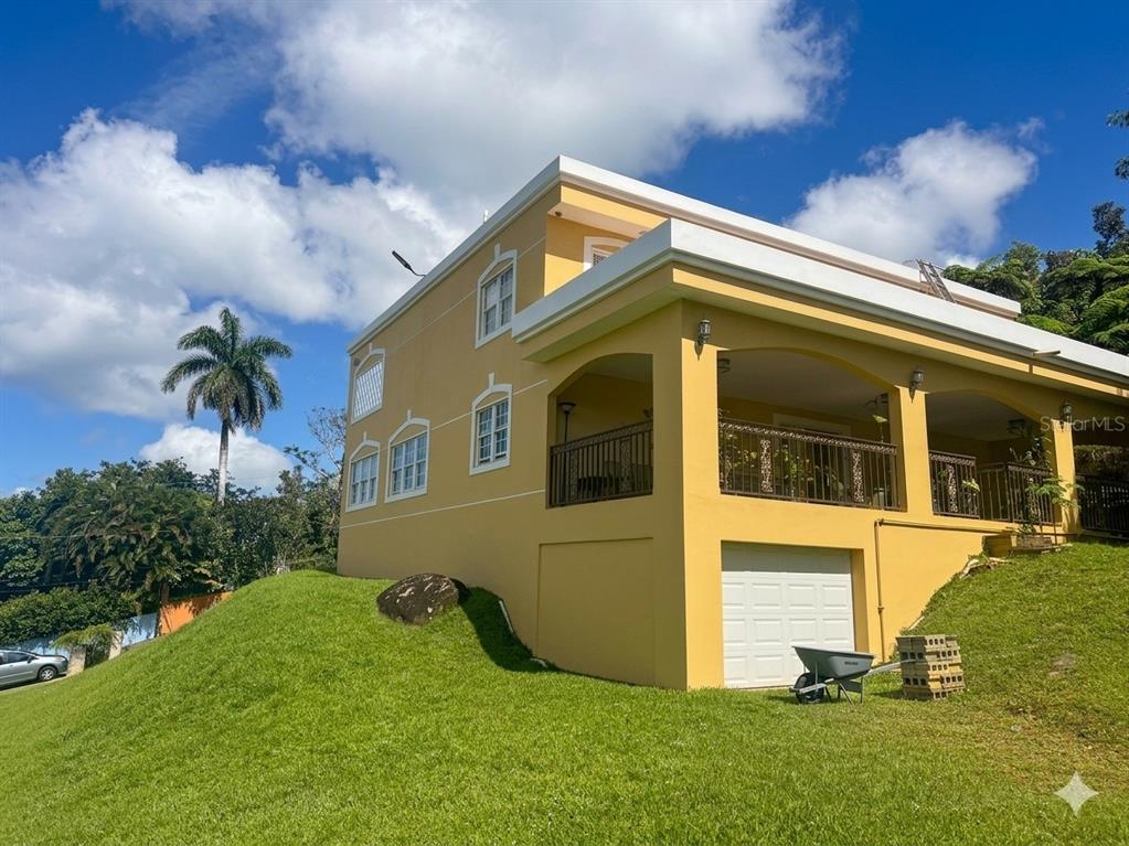 A yellow two-story house with a garage and a balcony sits on a grassy hill — Bo Beatriz Las Piñas FINCA MI RECREO  LOT 28, CAGUAS, PR