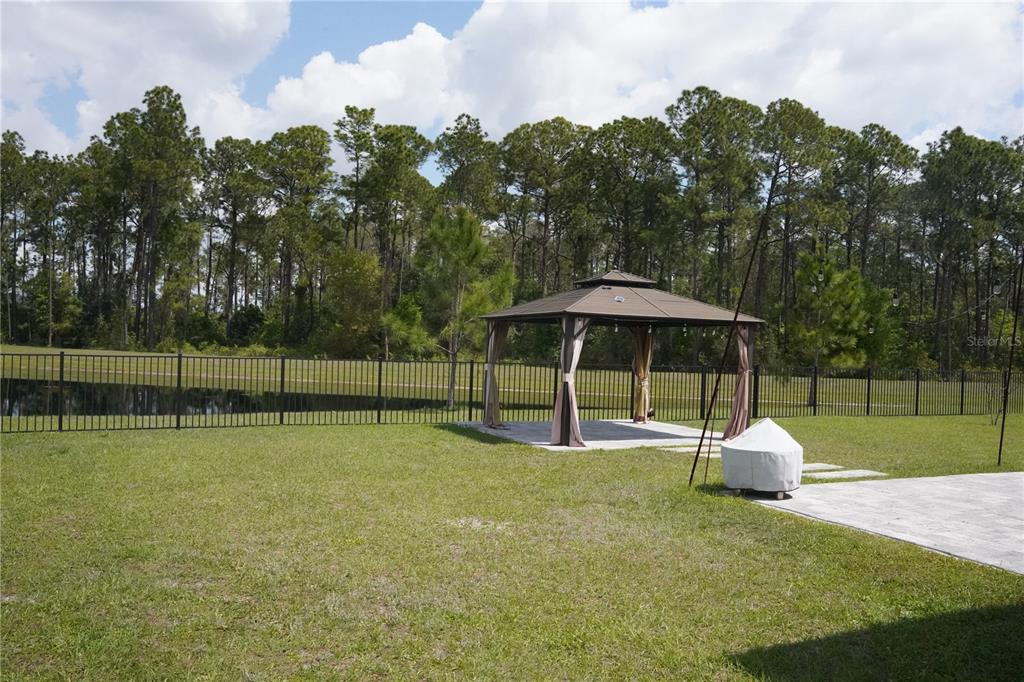 A backyard features a gazebo on a patio, a black metal fence, and a wooded background — 16436 WINDING PRESERVE CIRCLE, CLERMONT, FL